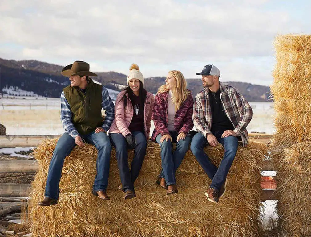 Four people are seated on a large, golden-brown hay bale in an outdoor setting.