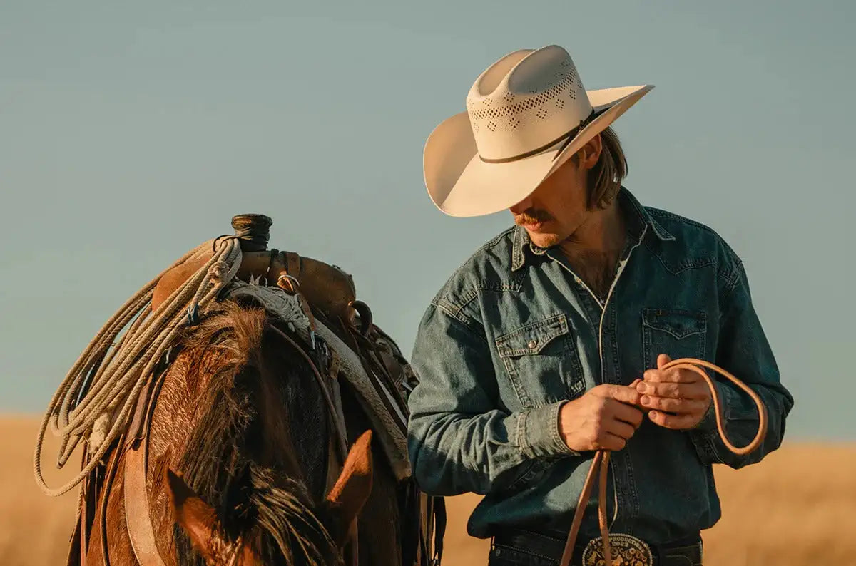 A man wearing a cream-colored cowboy hat with perforated detailing stands beside a horse, holding a leather rope.
