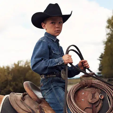 A young boy wearing a black cowboy hat and blue denim shirt, holding a coiled rope with leather straps.